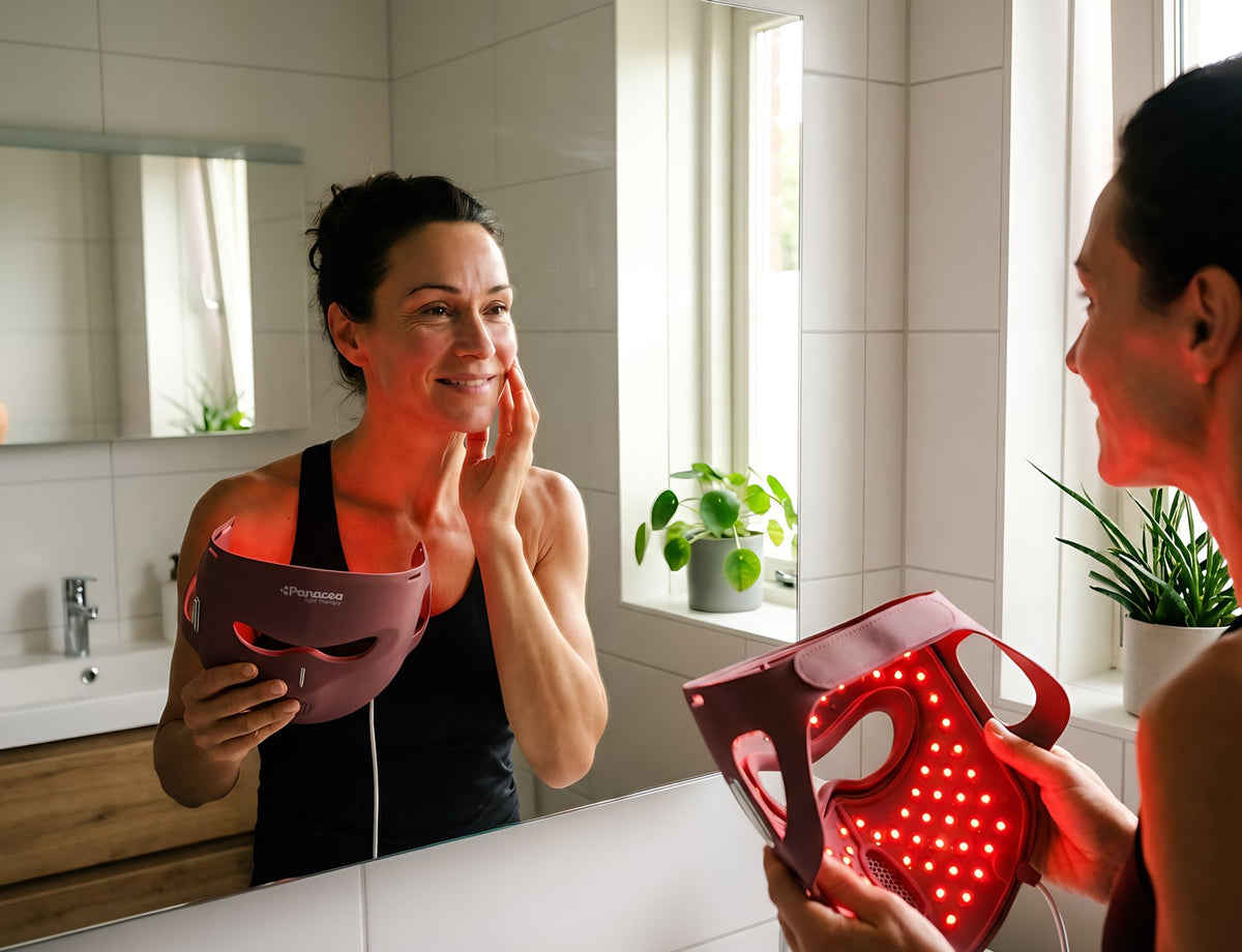 Woman looks at her skin in the bathroom mirror with the Panacea LED Mask in her hand